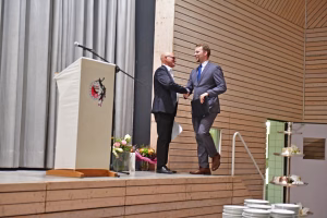 Zwei Männer im Anzug geben sich auf einer Bühne die Hand; im Hintergrund stehen Vorhang, Blumen und ein Banner als Anlassdekoration.