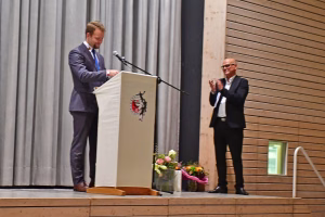 Redner am Podium mit Mikrofon, hält eine Ansprache; im Hintergrund steht ein weiterer Gast applaudierend neben Blumenarrangement und Tisch.