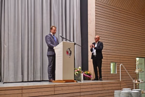 Zwei Redner stehen an einem Podium mit Mikrofon: eine Person hält eine Ansprache, die andere gestikuliert im Vortrag vor Publikum.