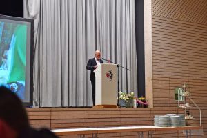 Mann spricht am Rednerpult vor einem Podium mit Mikrofon; dahinter Vorhänge und eine Projektion, daneben Deko-Blumen.
