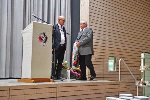 Zwei Männer stehen auf einer Bühne bei einer Rede; Rednerpult mit Mikrofon, Publikumshandlung, neben Blumenarrangements und gedecktem Tisch.