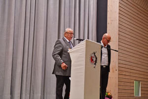 Redner am Podium mit Mikrofon, dahinter graue Vorhänge und Holzpanelen; eine weitere Person blickt von der Seite auf die Bühne.