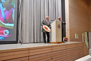 Zwei Redner stehen an einem Podium, halten eine Plakette und sprechen ins Mikrofon vor einem großen Vorhang; im Hintergrund eine Projektionsfläche.