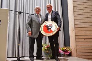 Zwei Männer stehen vor Vorhang und Mikrofon, halten gemeinsam einen runden Pokal als Auszeichnung bei einer feierlichen Sportveranstaltung.