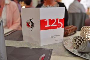 Weißes Jubiläumsgeschenk mit rotem „125“-Aufdruck auf einem Tisch, daneben Kerze im Glas und Deko-Teller.