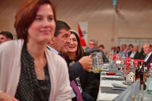 Teilnehmende bei einer Veranstaltung am gedeckten Tisch, im Vordergrund eine Frau im Gespräch, Hintergrund mit weiteren Gästen und Getränken.