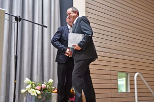 Gekleideter Mann umarmt und hält eine Urkunde, lächelt im Sitzen einer Veranstaltung; Blumenstrauß und Vorhang im Hintergrund.