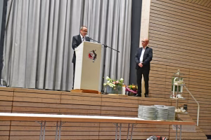 Zwei Personen stehen an einer Rednerpult-Bühne; eine spricht ins Mikrofon, die andere steht daneben. Blumenarrangement und Geschirr am Rand.