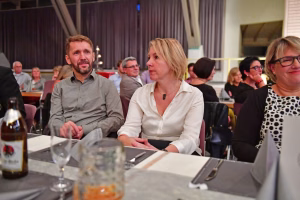 Zwei Personen sitzen an einem Tisch in einem Saal, sprechen und hören zu; Mikrofone und Technik deuten auf eine Veranstaltung oder Diskussion hin.
