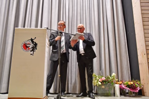 Zwei Männer am Rednerpult vor Vorhang und Mikrofon, einer hält eine Urkunde, der andere applaudiert; Blumen und Präsentiertisch im Vordergrund.