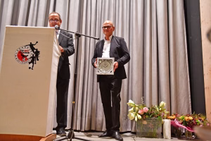 Zwei Männer stehen auf einer Bühne: einer spricht ans Mikrofon, der andere hält eine Trophäe; im Vordergrund Blumenarrangements.