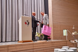 Zwei Männer am Rednerpult bei einer Veranstaltung; einer übergibt einem anderen ein pinkes Geschenk, im Hintergrund Vorhänge und Podium.