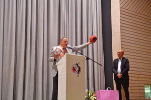 Mann bei einer Sportveranstaltung wirft einen Handball bei einer Ansage von der Bühne, neben ihm steht ein weiterer Mann.