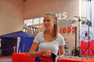 Porträt einer Person im Sportdress mit Blick in die Ferne, hält ein Gerät in der Hand; Hintergrund mit Arena-Bühne und Zelt.