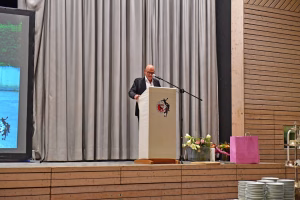 Redner an einem Podium mit Mikrofon, dahinter Vorhang; im Hintergrund Blumenarrangement und Geschirr deuten auf eine Veranstaltung.