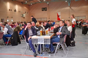 Teilnehmende sitzen an festlich gedeckten Tischen in einer Turnhalle, schauen nach vorn und hören einem Vortrag zu.