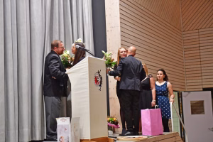 Personen auf einer Bühne bei einer Rede, Blumen und Mikrofone; eine Person überreicht/empfängt, rechts stehen weitere Gäste mit Geschenktüten.