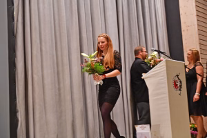 Junge Frau hält einen Blumenstrauß vor Vorhangkulisse bei einer Veranstaltung, während eine Person am Rednerpult im Hintergrund spricht.