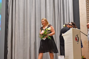 Frau mit Blumenstrauß und Kleid steht vor Vorhang, neben ihr spricht ein Mann hinter einem Podium mit Mikrofon.