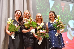 Vier lächelnde Frauen halten Blumensträuße vor einer Bühne mit Hintergrundbanner; sie stehen in Reihe für eine Ehrung/Feier.