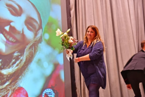 Person hält einen Blumenstrauß und steht vor einer Bühne; hinter ihr zeigt eine Projektion ein großes Bild, als Anlass einer Veranstaltung.