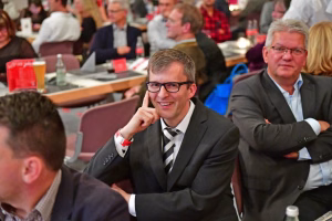 Zwei Männer in Anzügen bei einer Konferenzsitzung: einer stützt den Kopf, der andere sitzt abgewandt; sie wirken aufmerksam und wartend.