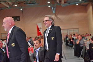 Zwei Männer im Anzug in einer Sporthalle, einer vor Zuschauern, der andere im Vordergrund; Blick und Gestik zeigen eine Ansprache.