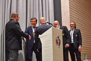 Mann überreicht am Rednerpult einem Publikum ein Präsent; mehrere Anwesende im Anzug lächeln, Mikrofon am Podium.