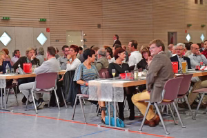 Gruppen sitzender und zuhörender Teilnehmender an festlich gedeckten Tischen in einer Sporthalle, im Hintergrund Veranstaltungssaal.