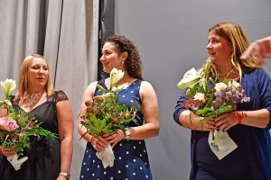 Drei Frauen halten Blumensträuße vor grauem Hintergrund; zwei lächeln, eine spricht begeistert—Bild zum Gratulieren bei einer Veranstaltung.