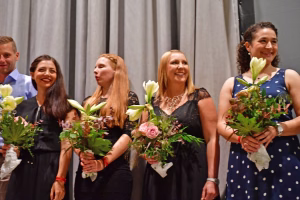 Vier Personen posieren mit Blumengestecken und lächelnden Gesichtern vor grauen Vorhängen; der Rahmen wirkt wie eine Feier oder Preisverleihung.