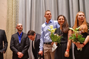 Mehrere Personen bei einer Feier, zwei stehen nebeneinander mit Blumen und blicken in die Kamera; eine Person verbeugt sich.