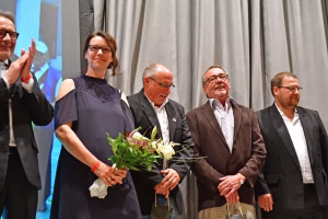 Personen bei einer Veranstaltung: Frau mit Blumenstrauß und lächelnden Gästen im Hintergrund, mehrere Männer im Anzug applaudieren.