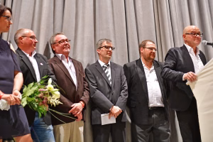 Mehrere Männer im Anzug sitzen auf einer Bühne; einer hält ein Mikrofon, andere applaudieren; im Vordergrund Blumenstrauß als Anlass.