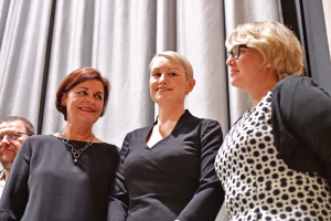 Drei Frauen in Businesskleidung bei einer sitzenden Versammlung, schauen nach vorn und lächeln; Gesprächs- oder Publikumsatmosphäre.