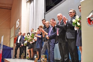 Mehrere Personen in Anzügen stehen auf einer Bühne, halten Blumen und applaudieren, während zwei Personen dem Publikum zuwinken und die Feier anführen.