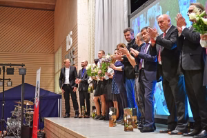 Festliche Bühne: mehrere Personen applaudieren und winken, einige halten Blumensträuße, vorn steht ein Podium mit Sektflaschen.