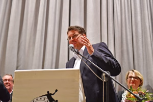 Redner hinter Pult mit Mikrofon, berührt sich am Gesicht und blickt in die Zuschauerrichtung vor beigem Bühnenvorhang.