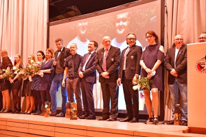 Gruppenfoto auf einer Bühne: Personen stehen im Halbkreis, einige halten Blumen, feiern einen Anlass bei Redebühne und Kerzenlichterdeko.