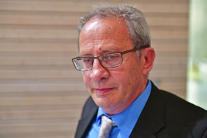 Nahaufnahme eines älteren Mannes mit Brille im Gespräch, ruhiger Gesichtsausdruck; unscharfer Hintergrund.