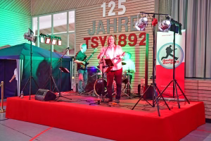 Rockband auf einer Bühne mit Gitarren und Mikrofonen, Scheinwerfern und großem Banner, präsentiert live in einem Indoor-Veranstaltungsraum.