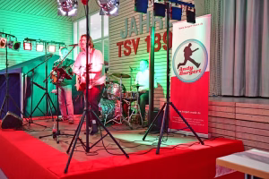 Musiker auf einer Bühne mit Mikrofon, Schlagzeug im Hintergrund; Scheinwerfer und Kabel, Banner rechts mit Gitarrensymbol zur Veranstaltung.