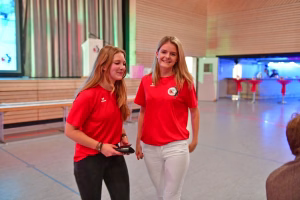 Zwei Personen in Sportkleidung stehen in einer Turnhalle, eine hält einen Ball, beide lächeln; Szene wirkt als Siegerfoto nach dem Spiel.