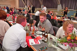 Festtafel in einer Sporthalle: mehrere Personen sitzen beisammen, unterhalten sich und trinken aus Gläsern. Getränke und Speisen stehen bereit.