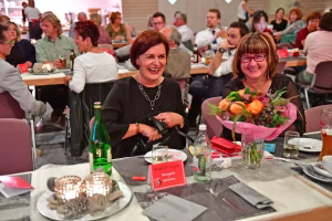 Zwei lachende Personen sitzen beim festlich gedeckten Tisch; im Vordergrund Blumengesteck und Geschirr, im Hintergrund weitere Gäste.