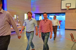 Zwei Männer laufen in einer Sporthalle auf die Kamera zu; im Hintergrund Basketballkorb und eine beleuchtete Veranstaltungsfläche.