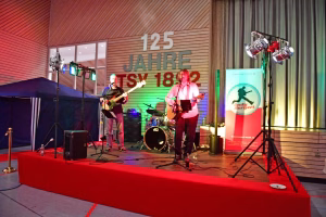 Live-Band auf einer Bühne mit zwei Gitarristen, Schlagzeug, Scheinwerfern und Mikrofonen vor großer Eventbeschriftung an der Wand.
