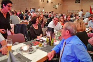 Geselliges Beisammensein bei einer Tischrunde in einem Saal: mehrere Personen sitzen beim Essen, Gespräche im Fokus.
