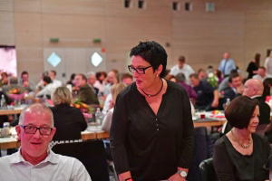 Teilnehmende beim Essen in einem großen Saal: Person mit Brille steht am Tisch, dahinter viele Gäste.