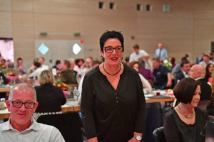 Lächelnde Frau mit Brille steht vorn im Saal; im Hintergrund sitzen viele Gäste bei einer Veranstaltung.
